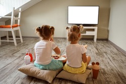 Two little girls are sitting on the floor on pillow, eating pizza and watching tv, playing games at home indoors. Two sisters spend time together, happy childhood.