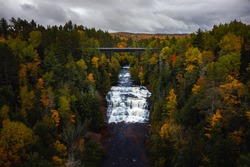 Aerial view of Agate Falls waterfall with an abandoned railroad bridge crossing above surrounded by evergreen and deciduous trees with fall colored foliage on a cloudy day in Upper Michigan.
