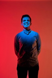 Fashion art portrait of smiling young guy face, holds hands behind his back. Red and blue light color.