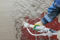 Plasterer operating sprayer equipment machine for spraying thin-layer putty plaster finishing on on a concrete wall covered with a red primer