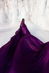 Art fantasy beautiful woman posing in a purple purple flying dress, with a long train of fabric. Behind her is a winter frozen shore covered in blue and white ice. The model stands with her back