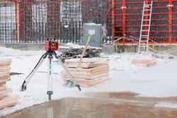 Geodetic device at snow covered construction site in winter season. Optical level at construction site. Equipment for work as surveyor. Optical theodelite on tripod. Red formwork on background