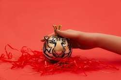A women's hand showing a decoration in the shape of a tiger on bright red background with shiny red tinsels around. Creative concept for Chinese New Year 2022 in the zodiac sign of the tiger. 