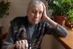 Portrait of a sad lonely old woman, sitting at home by the window. Old age, health problems, disabilities.	
