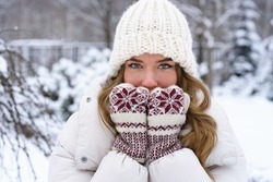 Portrait of a young woman in winter, outdoors. The beautiful woman covers her face with her hands in mittens against the cold. A very frosty, snowing winter day. 