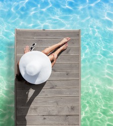 Summer holiday fashion concept - tanning woman wearing sun hat at the pool on a wooden pier view from above