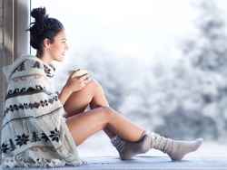 Young beautiful happy brunette woman drinking cup of coffee wearing knitted nordic print poncho sitting home by the window. Blurred winter snow tree background