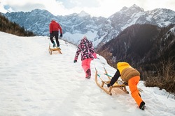 Father with two children in colorful snowsuits pulling wooden sleds uphill on a sunny winter day.