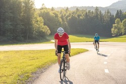 Female athlete on a racing bicycle riding uphill an asphalt road at sunset followed by a male cyclist.