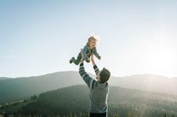 Portrait of a father holds, throws up happy daughter on hands walking on nature in autumn day. Dad and child playing in the mountains. Concept of family spending time together on vacation.
