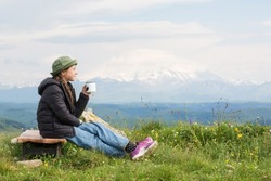 child teen sits against backdrop of mountains and drinks tea from cup in hike. travel, hiking, journey, outdoor recreation