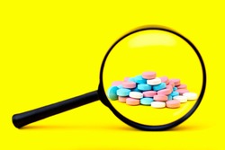 A look through a magnifying glass at a pile of multicolored pills on a yellow background. Search the ingredients of medicines. Identify fake and safe drugs in the pharmaceutical industry. Copy space.