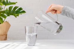 Woman hand with transparent jug fills a glass pure drinking water, creating air bubbles, green plant leaf on white table at home. Freshness, cold refreshing drink, water balance, health, thirst, diet.