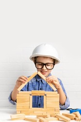 Smiling boy builder in a construction helmet builds a house from wooden blocks. Child engineer play with cubes at home. Mortgage, investment, choice of profession. Copy space.