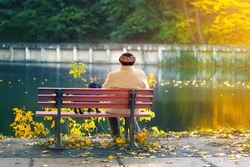 Lonely senior woman with a small black dog sitting on a bench by the autumn lake or river in a city park. Relaxation. Loneliness concept
