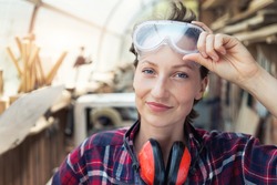 Young beautiful handy professional happy female strong carpenter portrait wearing protective goggles working in carpentry diy workshop against wood. Confident engineer. Women male hobby at workbench