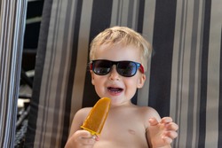 Portrait of little cute adorable caucasian blond little kid boy enjoy having fun relaxing and eat frozen fruit popsicle icecream sitting on sunbed yard near pool beach. Brother Child chilling outdoor