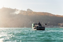 Scenic back view of fisherman angler enjoy fishing with rods on motor boat ship at clean green blue azure water harbor on early misty morning sunrise. Fishing speedboat with angling man at foggy bay