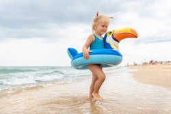 Portrait of cute little caucasian blond toddler baby girl in swimsuit and toucan inflatable ring toy enjoy swimming on sand sunny seaside beach on bright summer day. Family holiday vacation fun