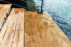 Close-up detail view of natural wooden stair steps of larch ladder into clean blue water of lake, sea or pond. Waterpoof timber material. Damp-proof striped decking board terrace surface