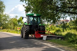 Big modern industrial tractor machine cutting green grass with mowing equipment along country roadside. Road lawn mower machinery vehicle highway maintenance service outdoors on sunny day