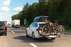 Back view modern white family wagon car with mounted roof box trunk and bike tail carrier driving european highway road against blue sky summer day. Lifestyle travel adventure trip journey concept