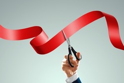 Grand opening with red ribbon and scissors. A businessman's hand holds scissors cuts a red ribbon on a light background. Close-up, copy space