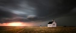 Old church in the rural countryside with a sever storm at sunset. There is an outhouse visible in the scene as well as a green and yellow grass meadow.