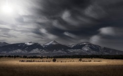Rocky Mountain landscape scene during the winter with snow-capped peaks and a meadow in the foreground with a dramatic dark sky and wispy white clouds.