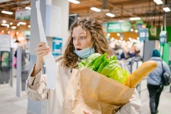 Young woman in a medical mask looks shocked at a paper check in a grocery supermarket holding a paper bag with groceries, price increase and inflation