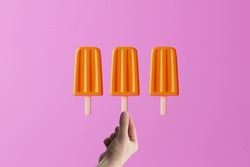 Hand holds orange ice lolly on an pink background. Concept of summer, vacation. Cooling down on warm days. 
