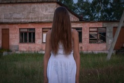 girl in a white dress with long hair thrown over her face stands in the twilight against the background of an old abandoned building, a school. concept of horror, mysticism