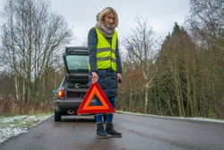 Young blonde woman in a winter down jacket in a yellow vest holds an emergency stop sign an orange triangle near a car with an open trunk