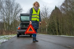 Young blonde woman in a winter down jacket in a yellow vest holds an emergency stop sign an orange triangle near a car with an open trunk