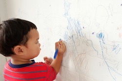 Cute asian boy in bright red striped t-shirt stands scribbling on white wall. with text copy space