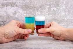 hands hold glasses with cocktails in the colors of the flags of Ukraine and Russia and clink glasses