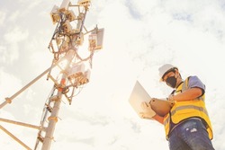 Male engineer wearing a mask working outdoors uses a laptop for maintenance to control the installation and inspection of the telecom tower network communication technology network.