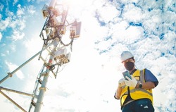 Male engineer uses a smartphone to connect to a 5G network,working in the field near a telecommunication tower that controls cellular electrical installations : Telecom modern communication technology