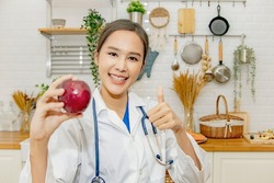 Vibrant and healthy beautiful asian woman nutritionist doctor thumbs up showing red apple, miracle fruit with vitamin C, excellent nutrition, good for health, nutrition advice in the kitchen.