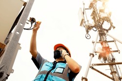 Male electrical engineer wearing helmet and mask uses screwdriver check the safety sensor connector outside the telecommunication tower control cabinet and uses smartphone the maintenance team reports