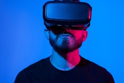 Young bearded man in virtual reality helmet plays online game in metaverse. Game simulates behavior in fictional world, gadgets and virtual reality addiction. Future technology concept.