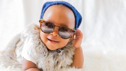 Super cool baby. Hipster baby in fur vest and sunglasses lies on a white bed in a room with curtains. Baby smiles while putting on sunglasses. 