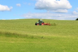 Farmer with agricultural equipment mowing the meadow and making hay. Hilly foothills of the Alps, blue sky, Allgäu, Bavaria.