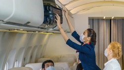 asian air hostess helping senior female caucasian passenger putting luggage into luggage cabin in airplane