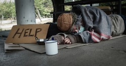 Homeless man lies on the ground while holding his mug and ask for help. He is unemployed and hungry due to the Coronavirus contamination.