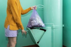 A woman in casual clothes throws a bag of garbage into the garbage chute