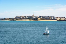 General view of the walled city of Saint Malo in Brittany, France, with the steeple of the cathedral protruding above the buildings behind the wall and a boat sailing on the sea in the foreground.