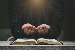 Man hands praying to god with the bible. Yellow lights and sparkles coming on Believe in goodness. Holding hands in prayer on a wooden table. Power of hope or love and devotion. 