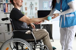 Close-up of medical worker helping to develop arm lifting blue dumbbell. Nurse in uniform working with disabled person. Man in wheelchair in clinic. Medicine and physical therapist concept