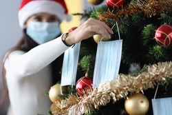 Woman in santa claus hat hanging protective medical face mask on christmas tree. Celebrating new year at home during covid-19 pandemic concept